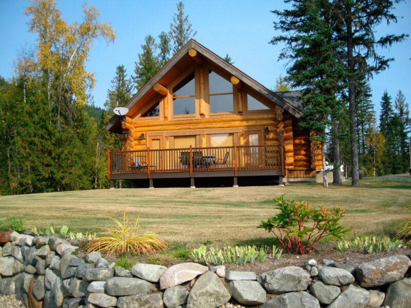 A log cabin with a stone wall in the foreground with trees.